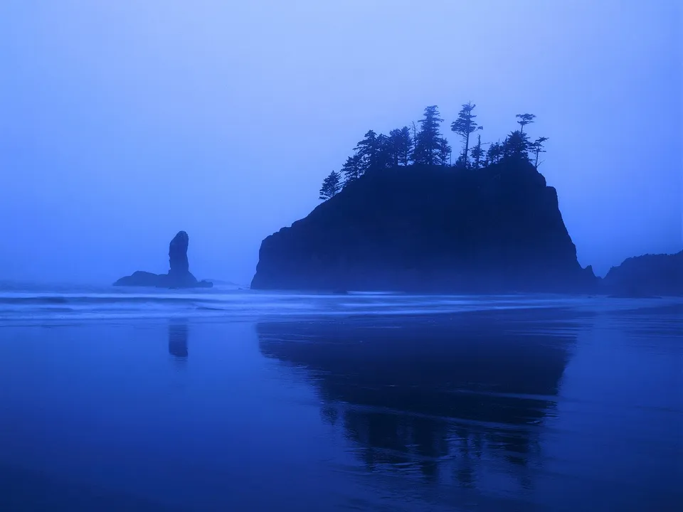A dramatic sea stack stands tall against a misty blue sky, with waves lapping the shore and reflections on wet sand.