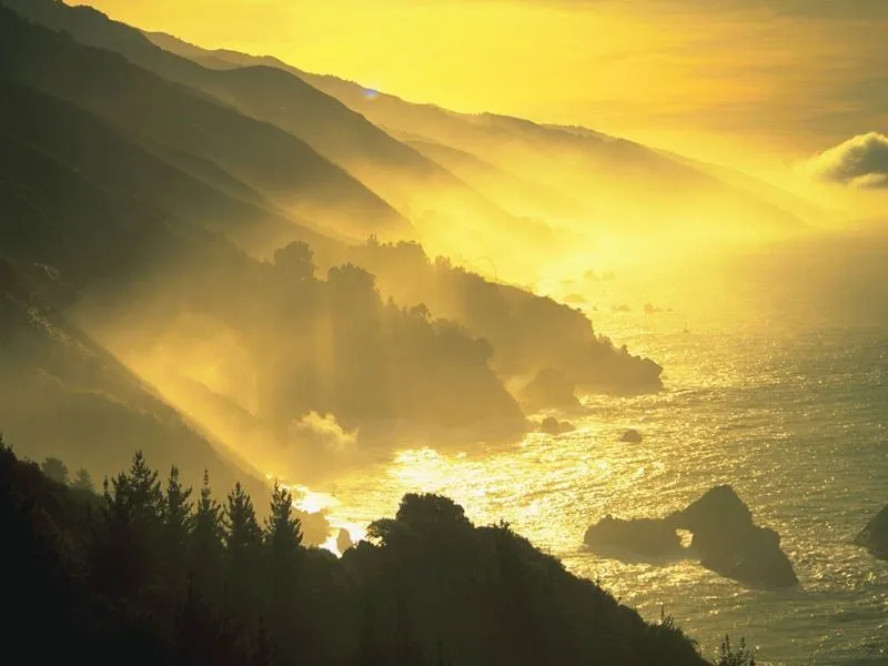 Sunlight streams through mist over a rugged coastline with waves crashing against rocks.