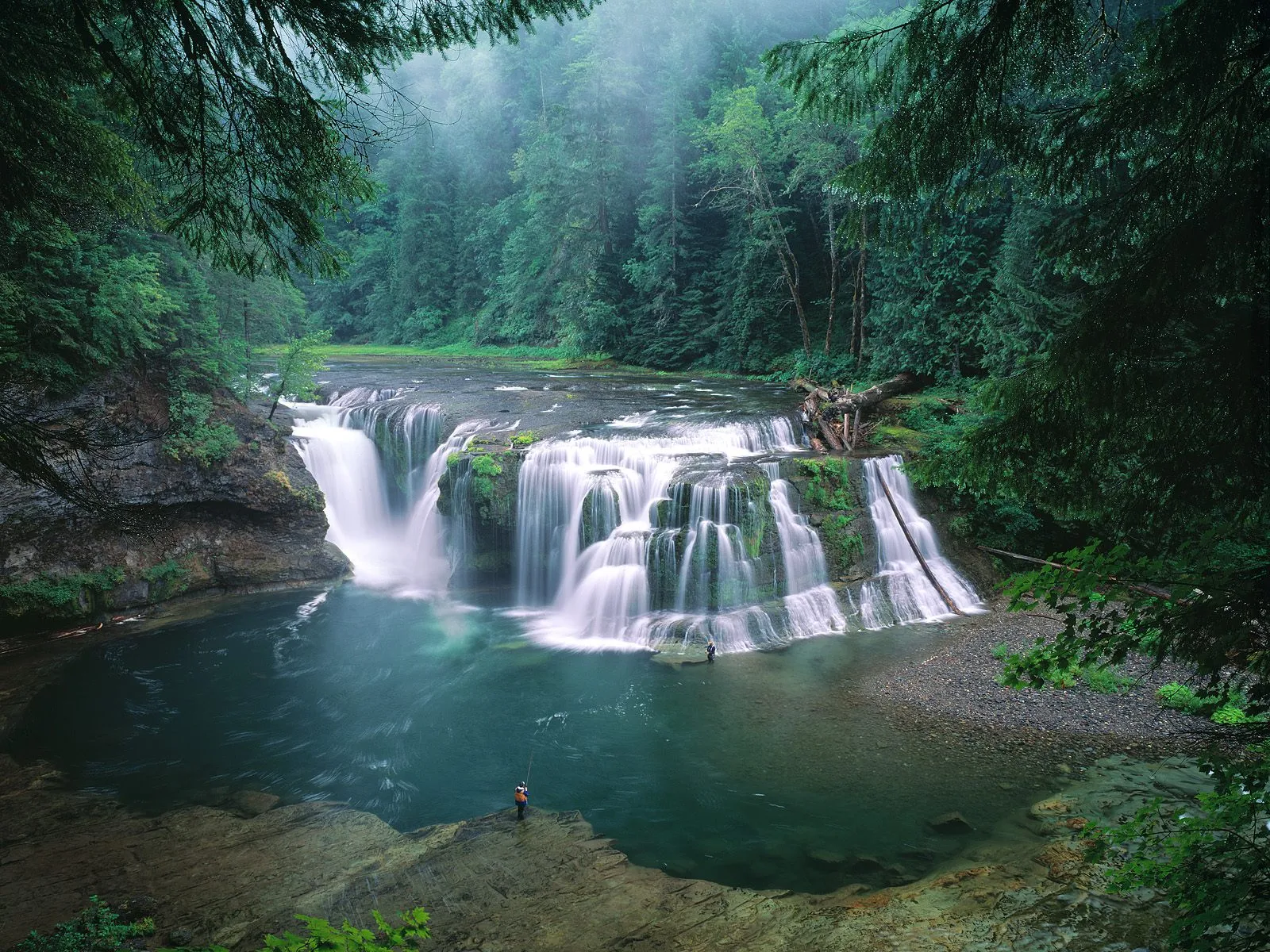 A wide, multi-tiered waterfall cascades into a turquoise pool, surrounded by lush green forest and a lone angler.