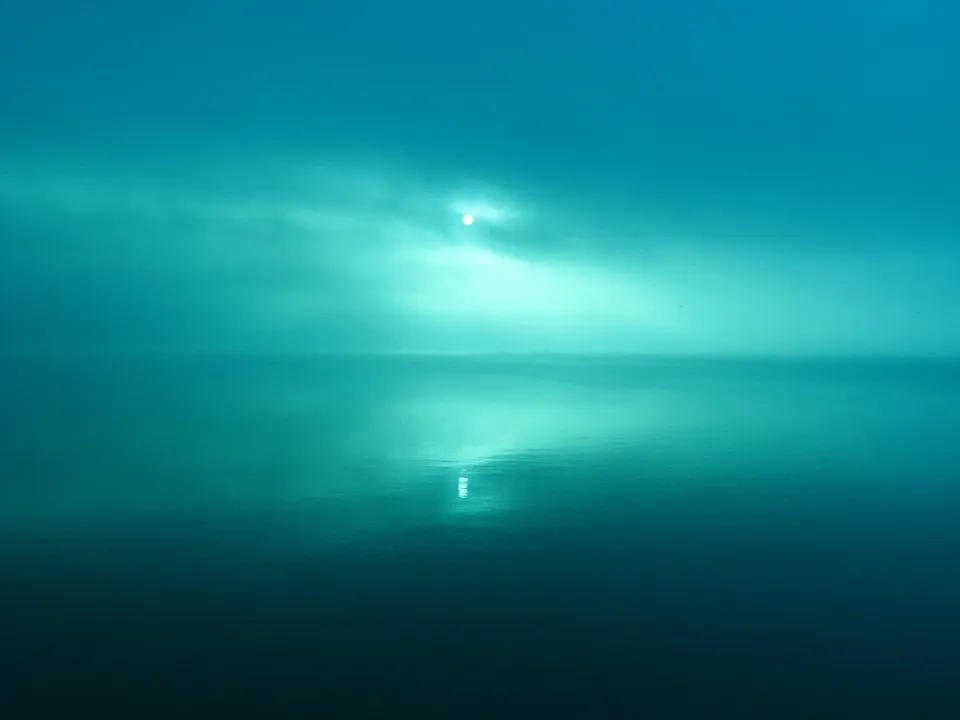 A hazy sun peeks through clouds over a vast, calm body of water with a reflection.