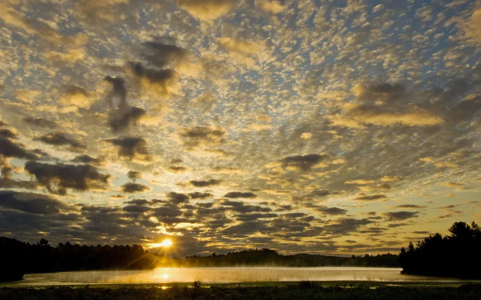 The sun rises over a misty lake, casting golden light through scattered clouds.