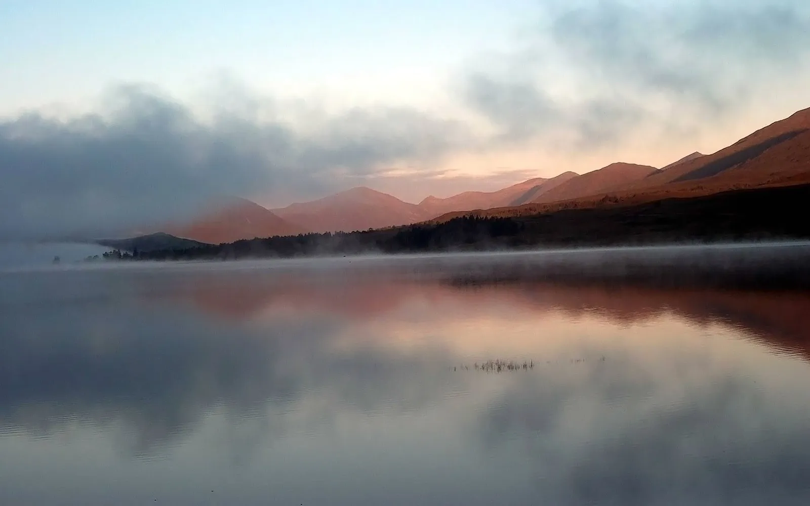 A misty lake with mountains in the background, reflecting the soft colors of sunrise.