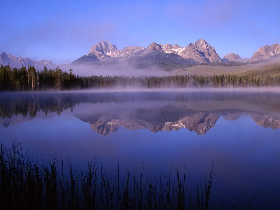 A serene mountain lake reflects jagged peaks and a line of pine trees through morning mist.