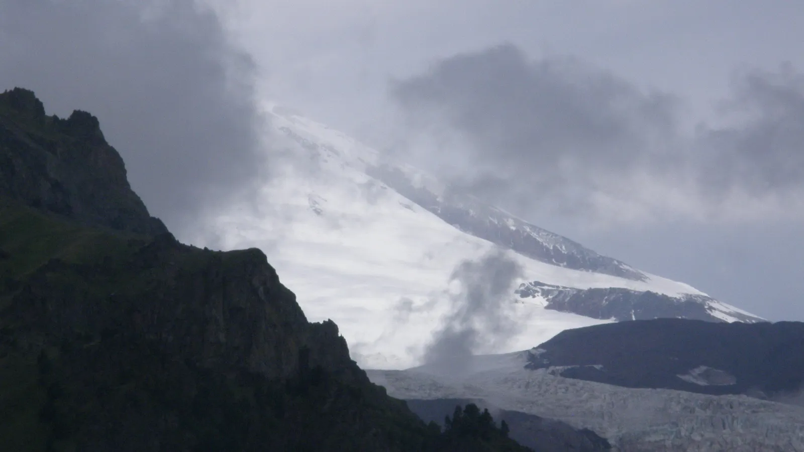 A dark, rocky mountain peak is in the foreground, with a snow-covered mountain range shrouded in mist and clouds in the background.
