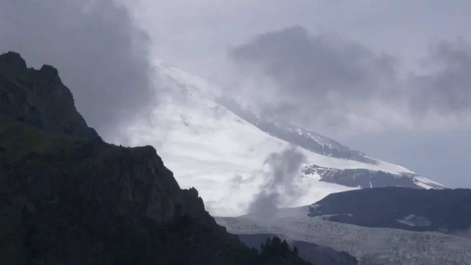 A dark, rocky mountain peak is in the foreground, with a snow-covered mountain range shrouded in mist and clouds in the background.