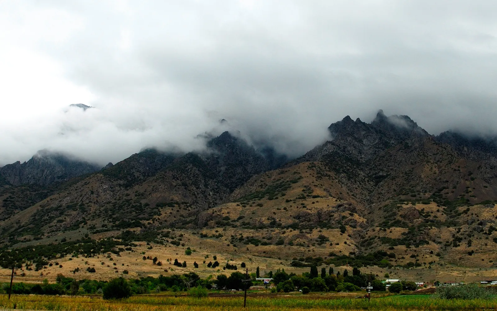 A rugged mountain range is partially obscured by low-hanging clouds, with a valley and trees in the foreground.