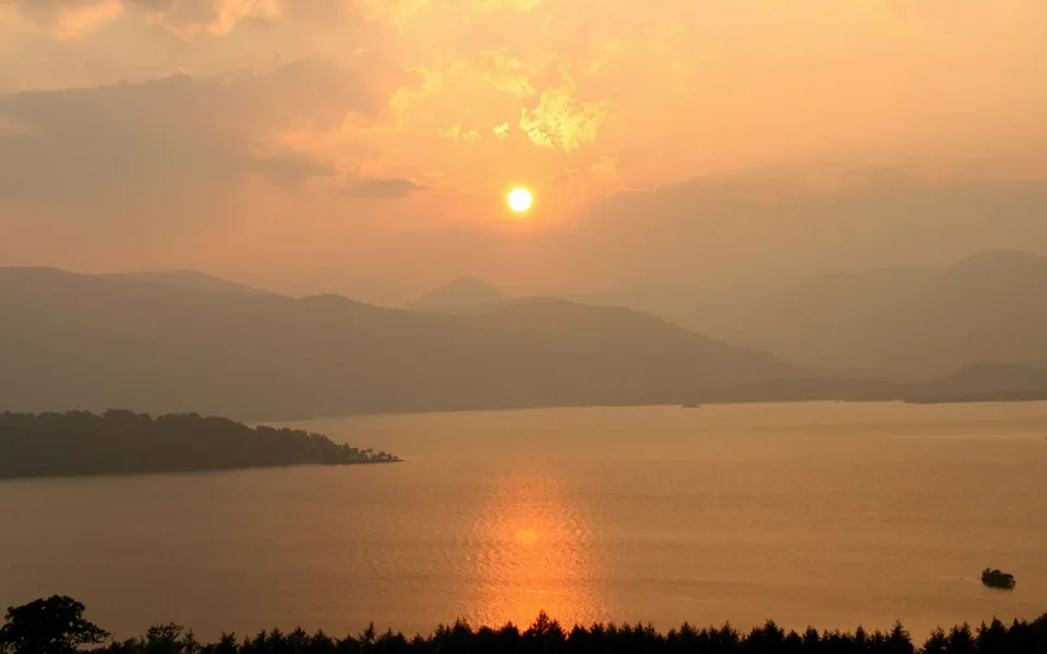A hazy sunset over a wide lake with silhouetted mountains in the background.