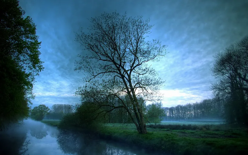 A calm river flows through a misty landscape with trees on either side and a dramatic cloudy sky.