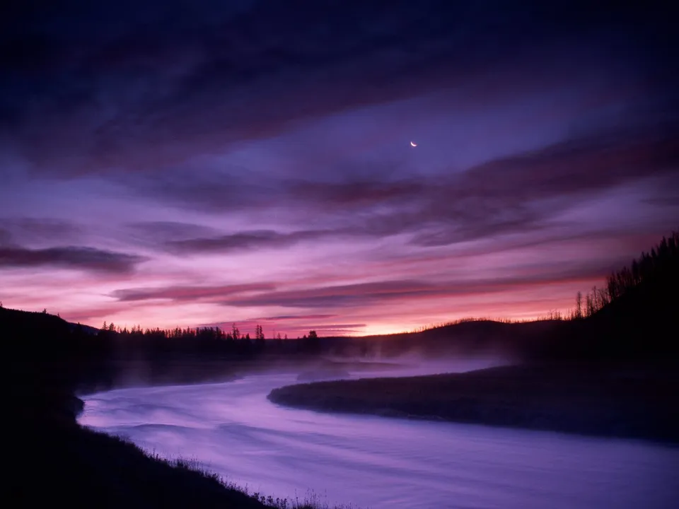 A winding river flows through a misty landscape under a dramatic purple and pink sky with a crescent moon.