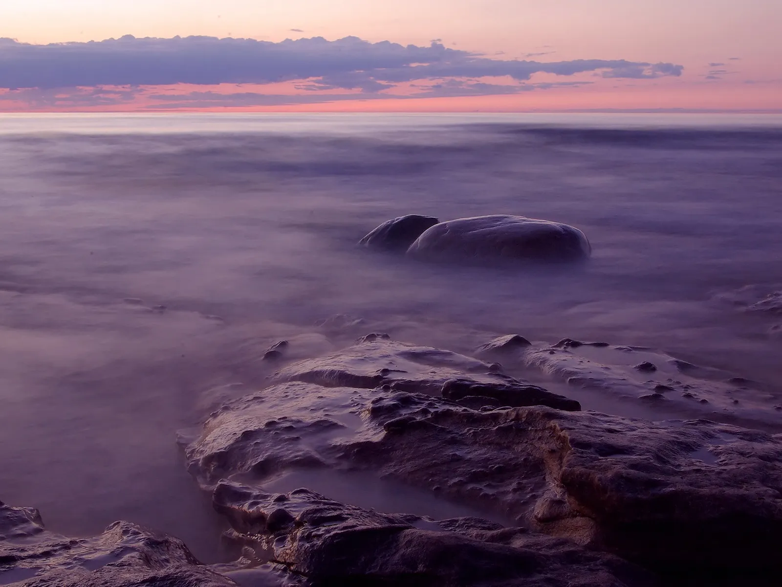 Long exposure shot of a rocky shoreline with smooth water and a pastel sunset sky.