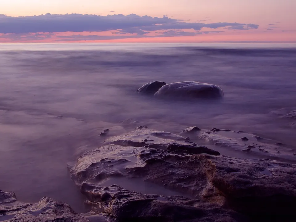 Long exposure shot of a rocky shoreline with smooth water and a pastel sunset sky.