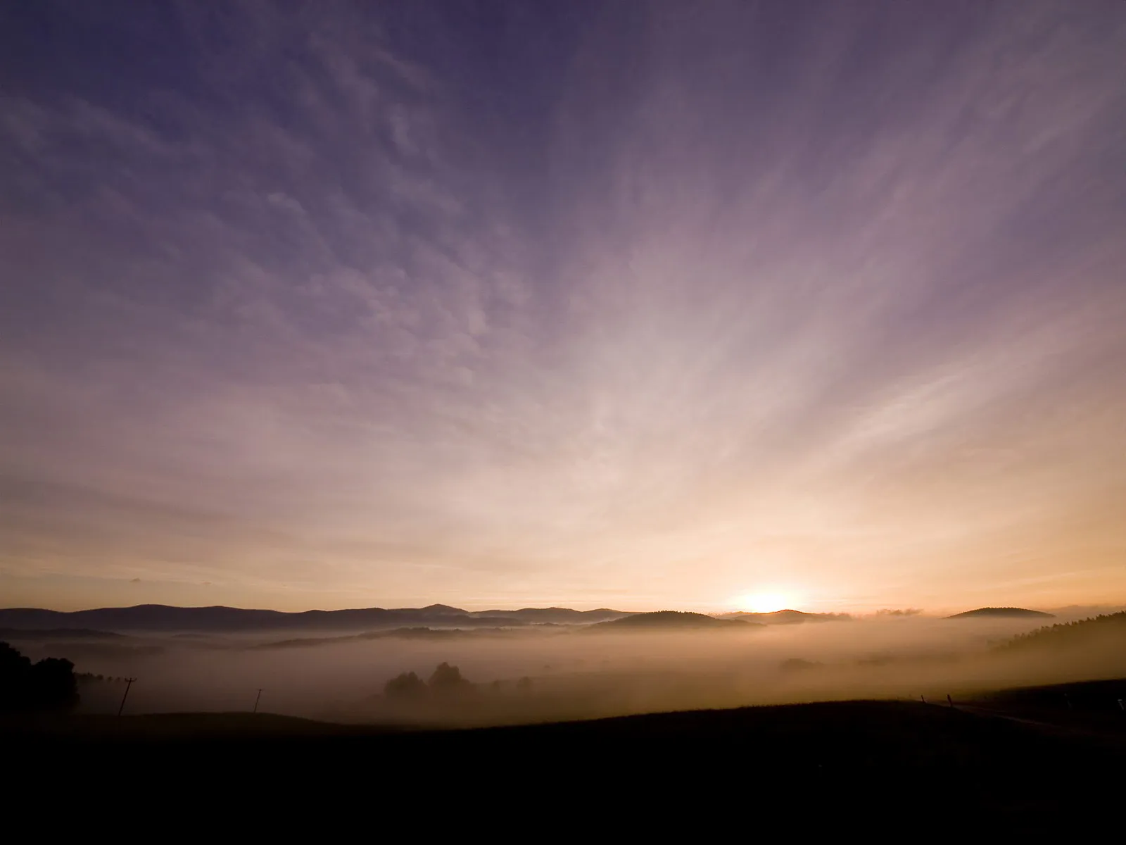 A valley filled with mist is illuminated by the golden light of a sunrise, with rolling hills in the background.