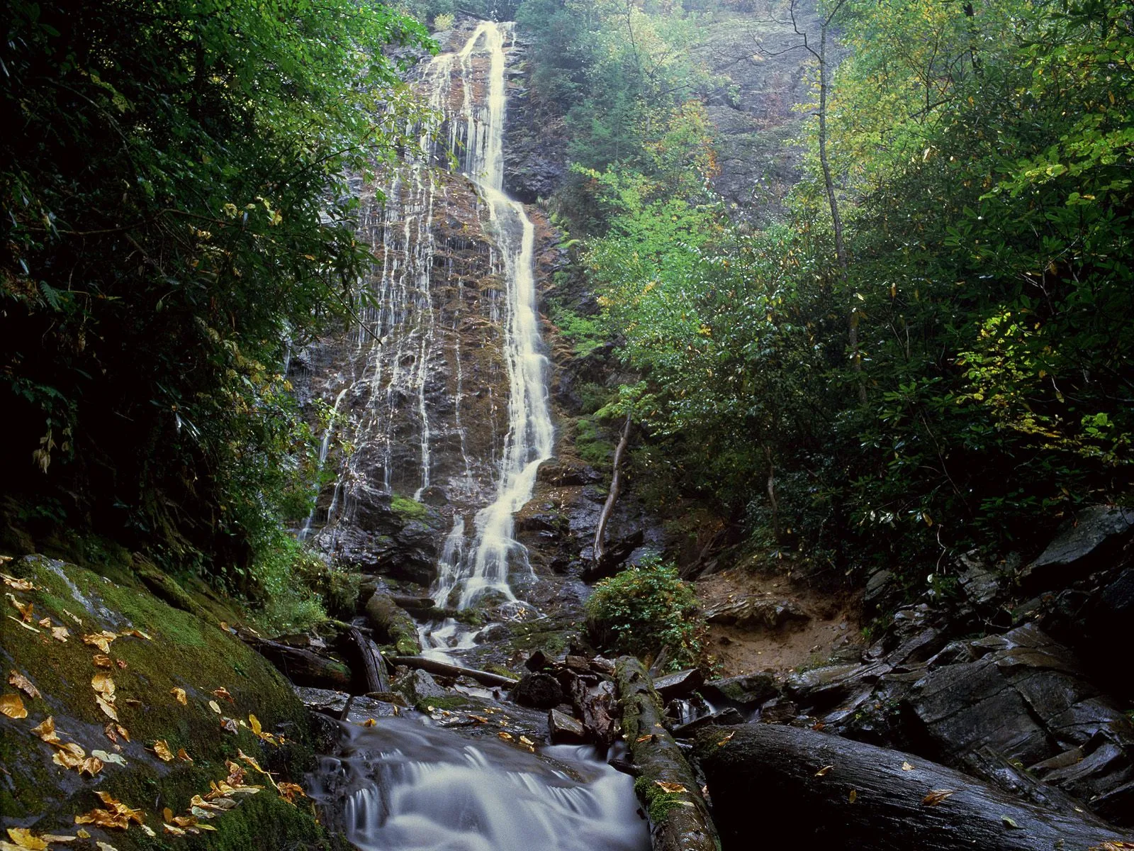 A tall, slender waterfall flows down moss-covered rocks, appearing misty due to a long exposure.