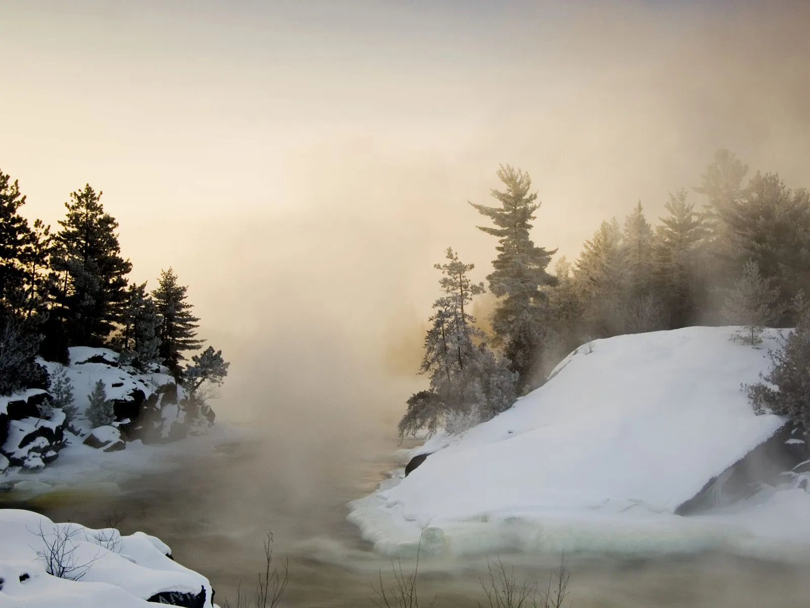 A misty winter scene with snow-covered banks and trees along a flowing river.