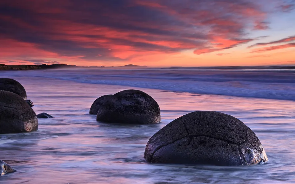 Large spherical boulders sit in the ocean water at sunset with a vibrant pink and orange sky.
