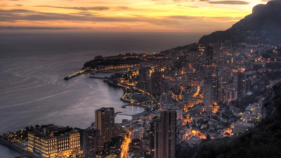 A wide aerial view of Monaco at dusk, with city lights twinkling and the Mediterranean Sea stretching to the horizon.