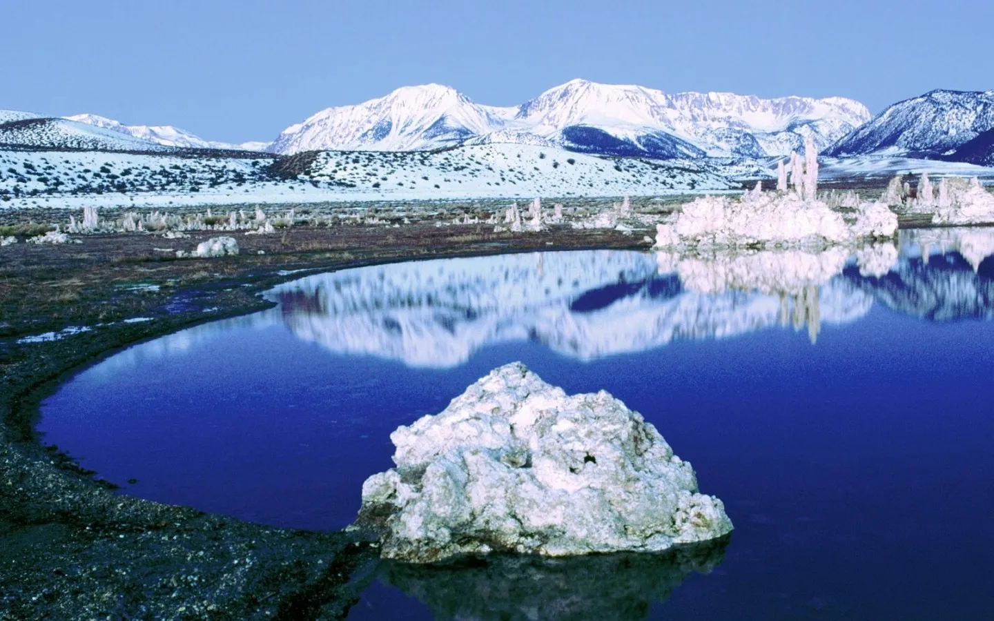 A calm lake reflects snow-capped mountains and white tufa formations under a clear blue sky.