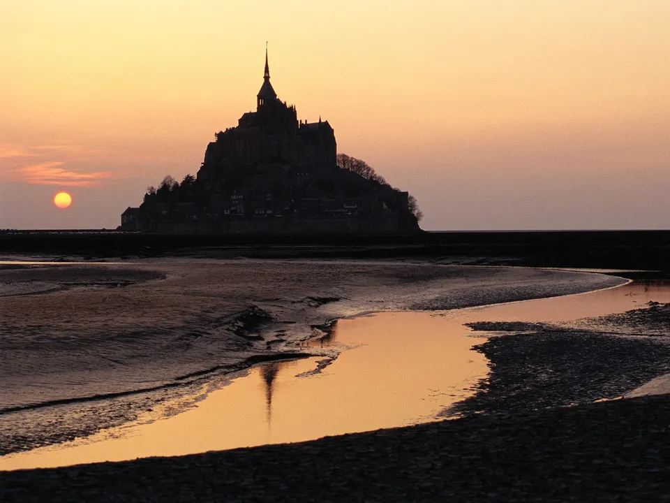 A silhouette of Mont Saint-Michel castle on a tidal flat during sunset, with the sun low on the horizon and reflections in the water.