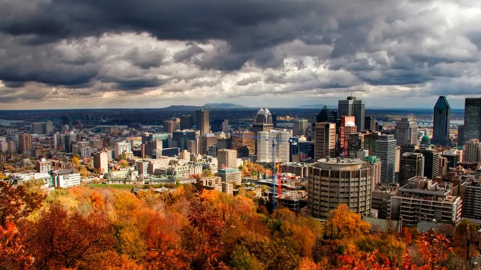 A panoramic view of the Montreal skyline with vibrant autumn foliage in the foreground and dark, dramatic clouds overhead.