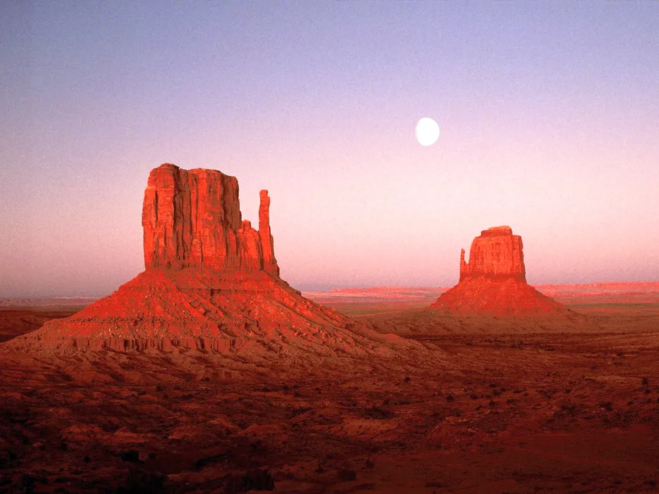 Two large sandstone buttes stand in Monument Valley, illuminated by the soft light of sunset.