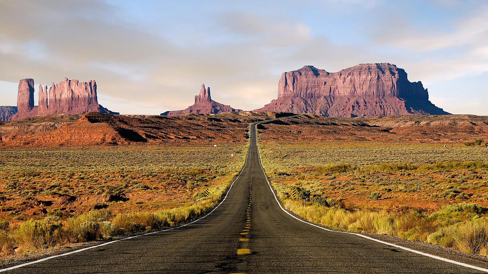 A long, empty asphalt road stretches towards the iconic red rock formations of Monument Valley under a partly cloudy sky.