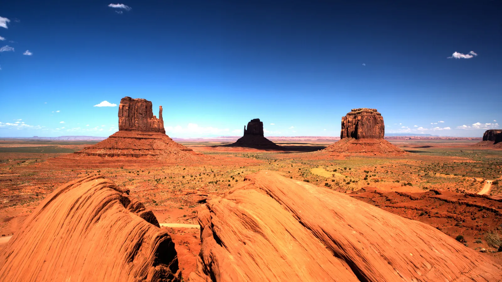 Three large sandstone buttes stand in Monument Valley under a bright blue sky with scattered clouds.