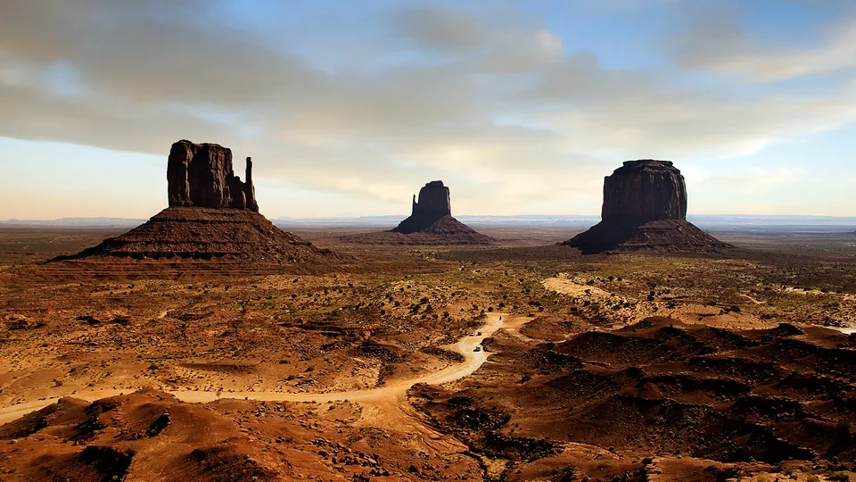 Three large sandstone buttes, known as the Mittens, stand in Monument Valley under a partly cloudy sky.