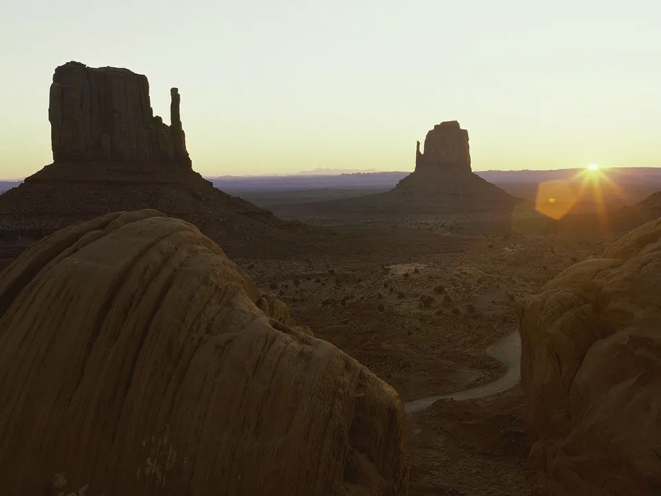 The sun rises over Monument Valley, creating a sun flare between the Mittens buttes.