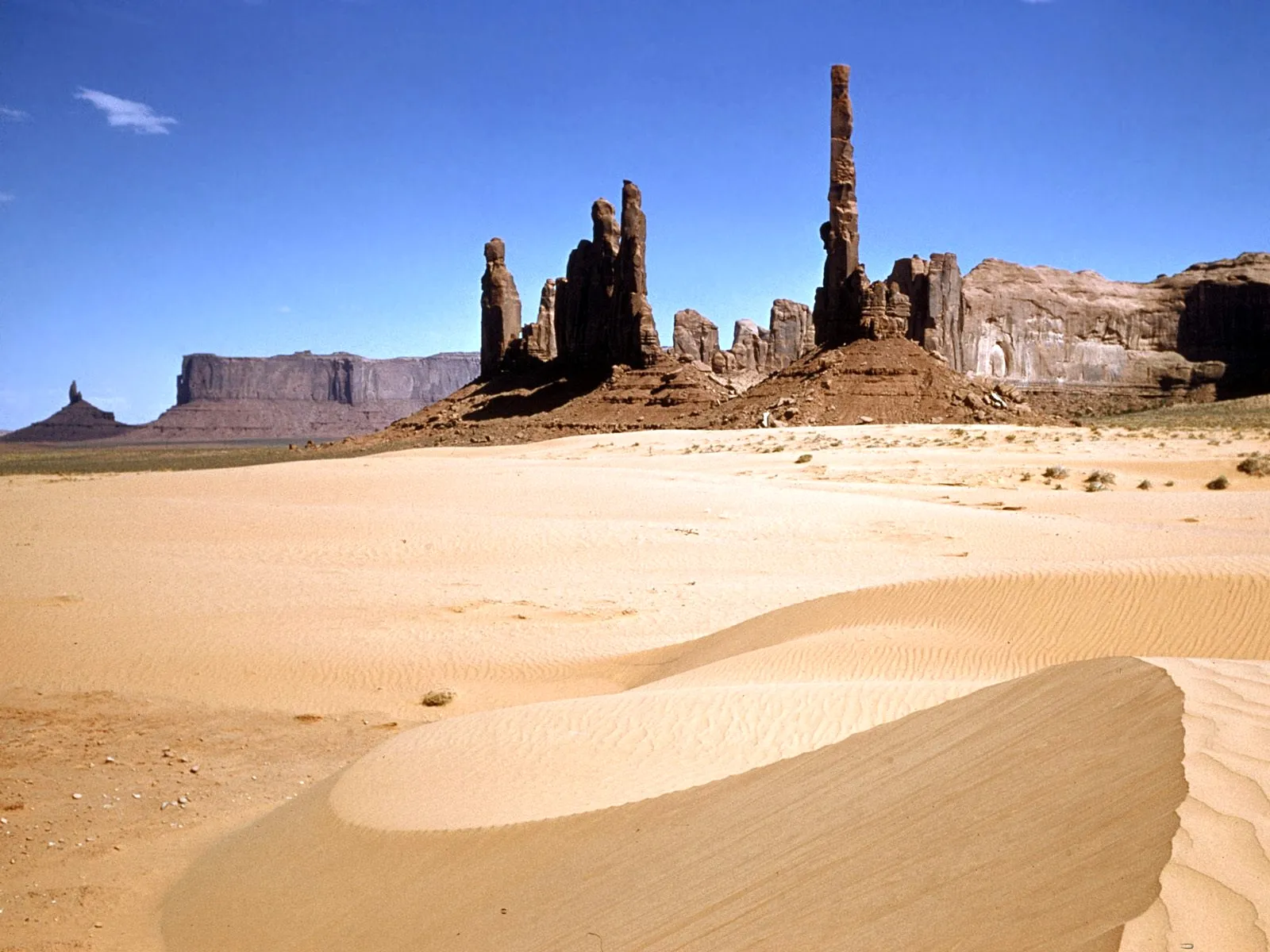 Rolling sand dunes in Monument Valley with dramatic rock formations and a clear blue sky.