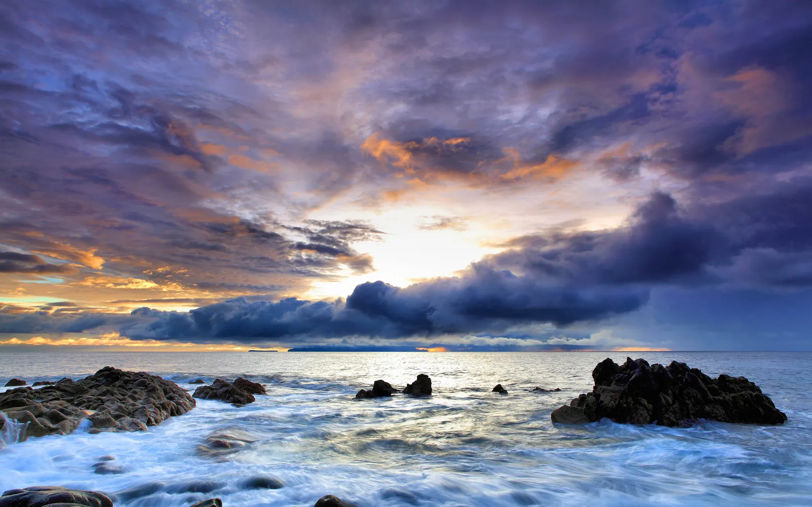 Dramatic clouds in purple, blue, and orange fill the sky as waves crash against dark, rugged rocks.