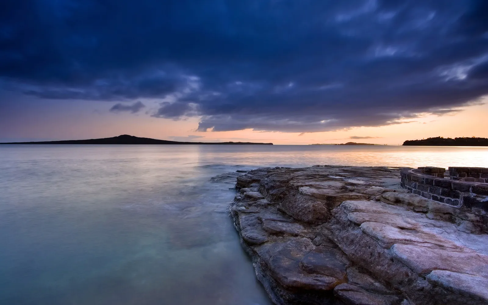 A moody sunset over calm water with a distant island silhouette and rocky foreground.