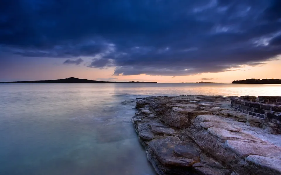 A moody sunset over calm water with a distant island silhouette and rocky foreground.