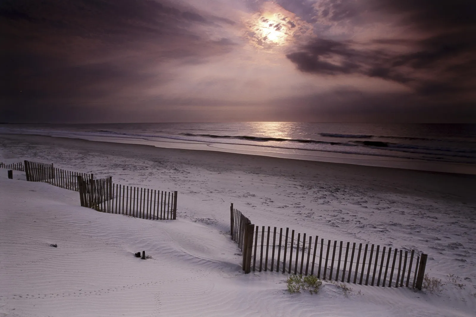 A sandy beach with a wooden fence in the foreground under a cloudy sky with the moon shining through.