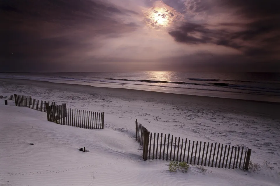 A sandy beach with a wooden fence in the foreground under a cloudy sky with the moon shining through.