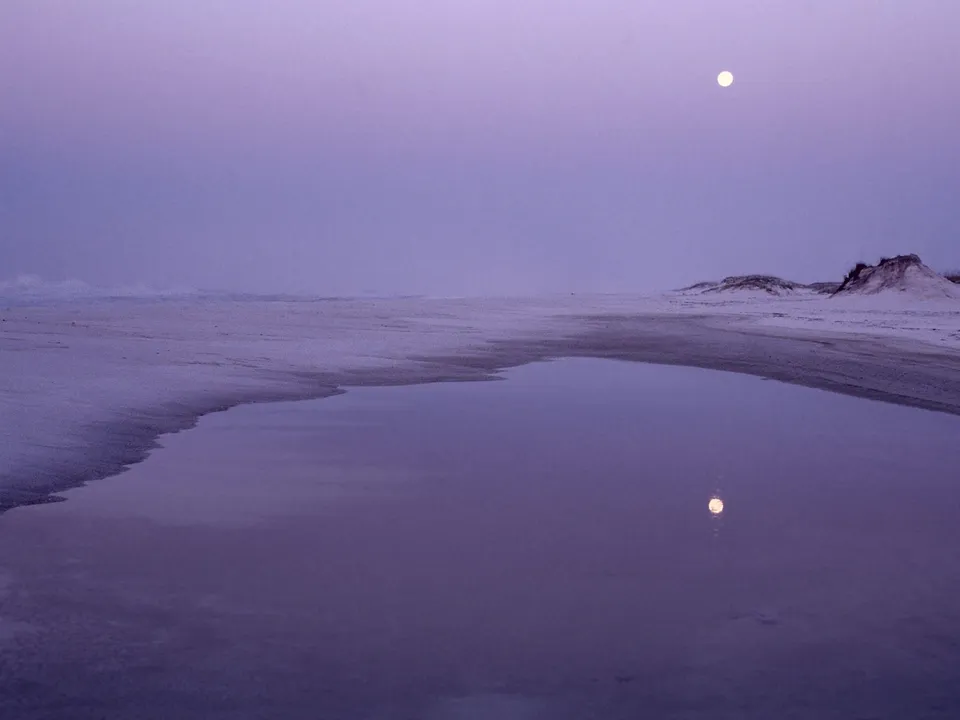 A serene beach at dusk with a full moon reflected in a calm tide pool.