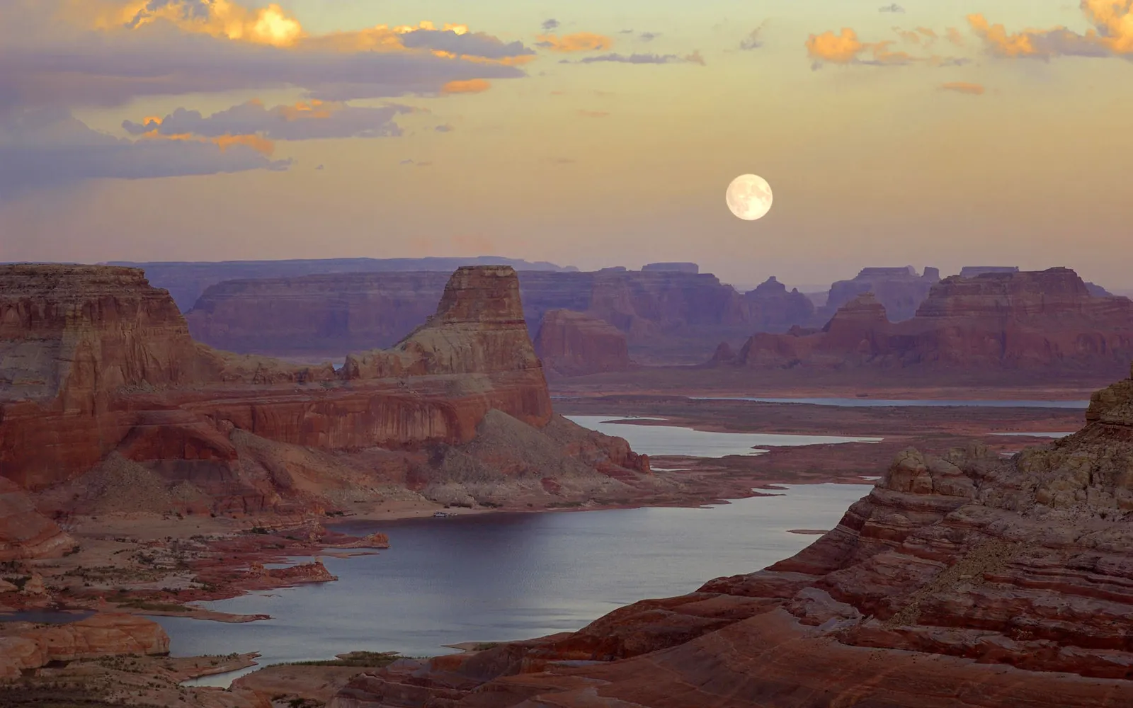 A full moon hangs in the sky over a vast canyon landscape with a winding river.