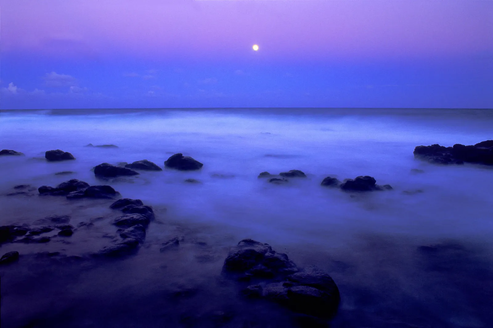 A full moon shines in a purple sky over a rocky coastline with misty ocean waves.