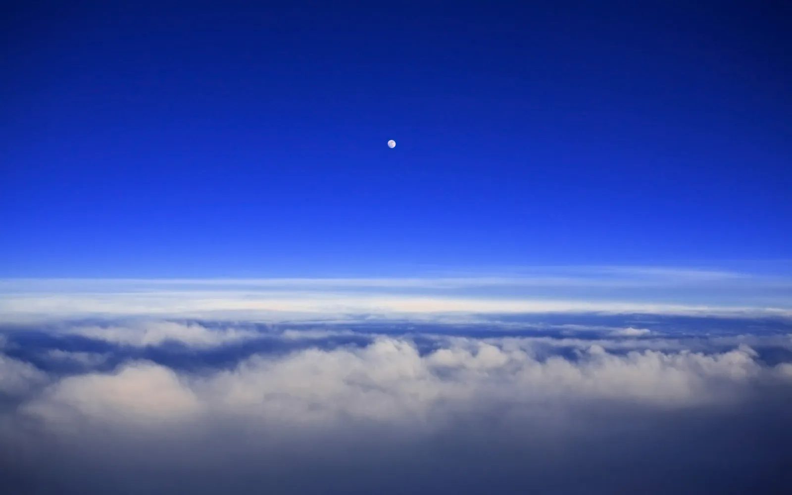A small, bright moon is visible in a deep blue sky above a thick layer of white and gray clouds.
