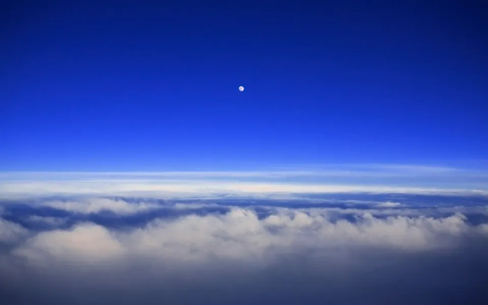 A small, bright moon is visible in a deep blue sky above a thick layer of white and gray clouds.