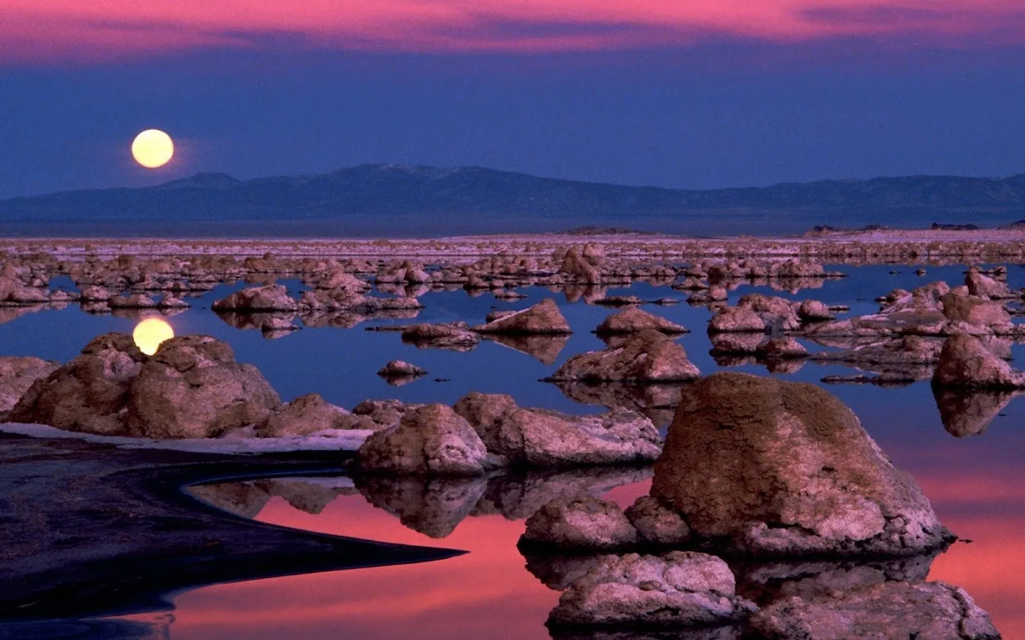 A full moon shines over a lake dotted with tufa rock formations, reflecting the pink and blue hues of the sky.