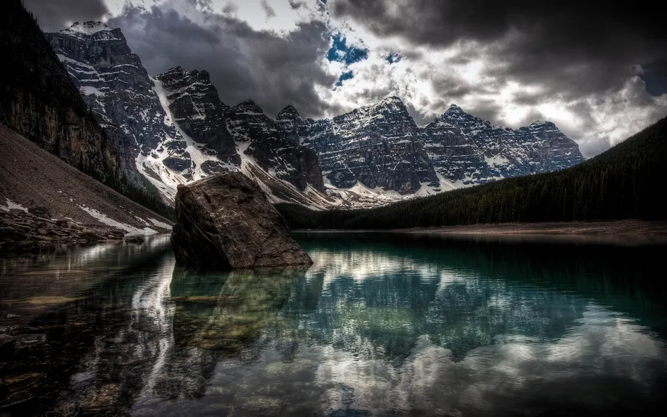 A large boulder sits in the turquoise waters of Moraine Lake, reflecting the dramatic, snow-dusted mountains and cloudy sky.