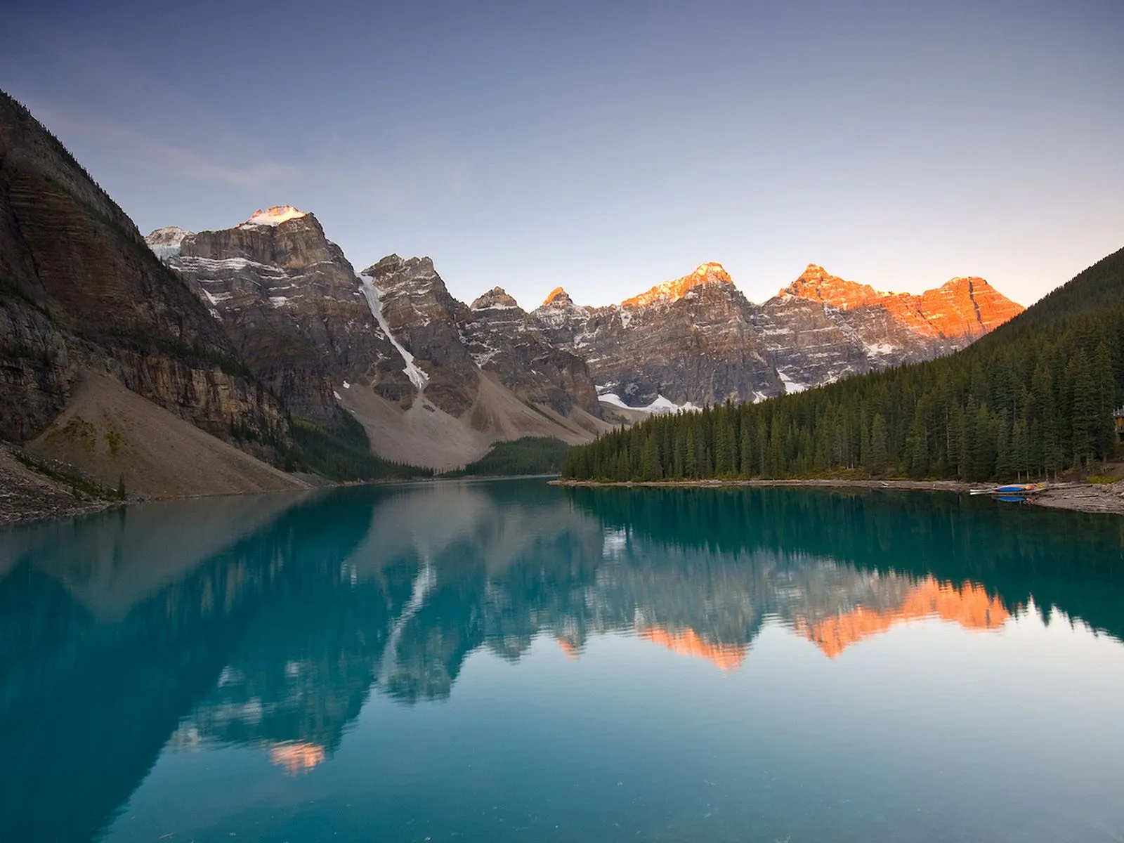 A stunning turquoise lake reflects snow-capped mountains and a pine forest under a clear sky at sunrise.