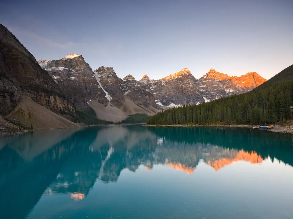 A stunning turquoise lake reflects snow-capped mountains and a pine forest under a clear sky at sunrise.
