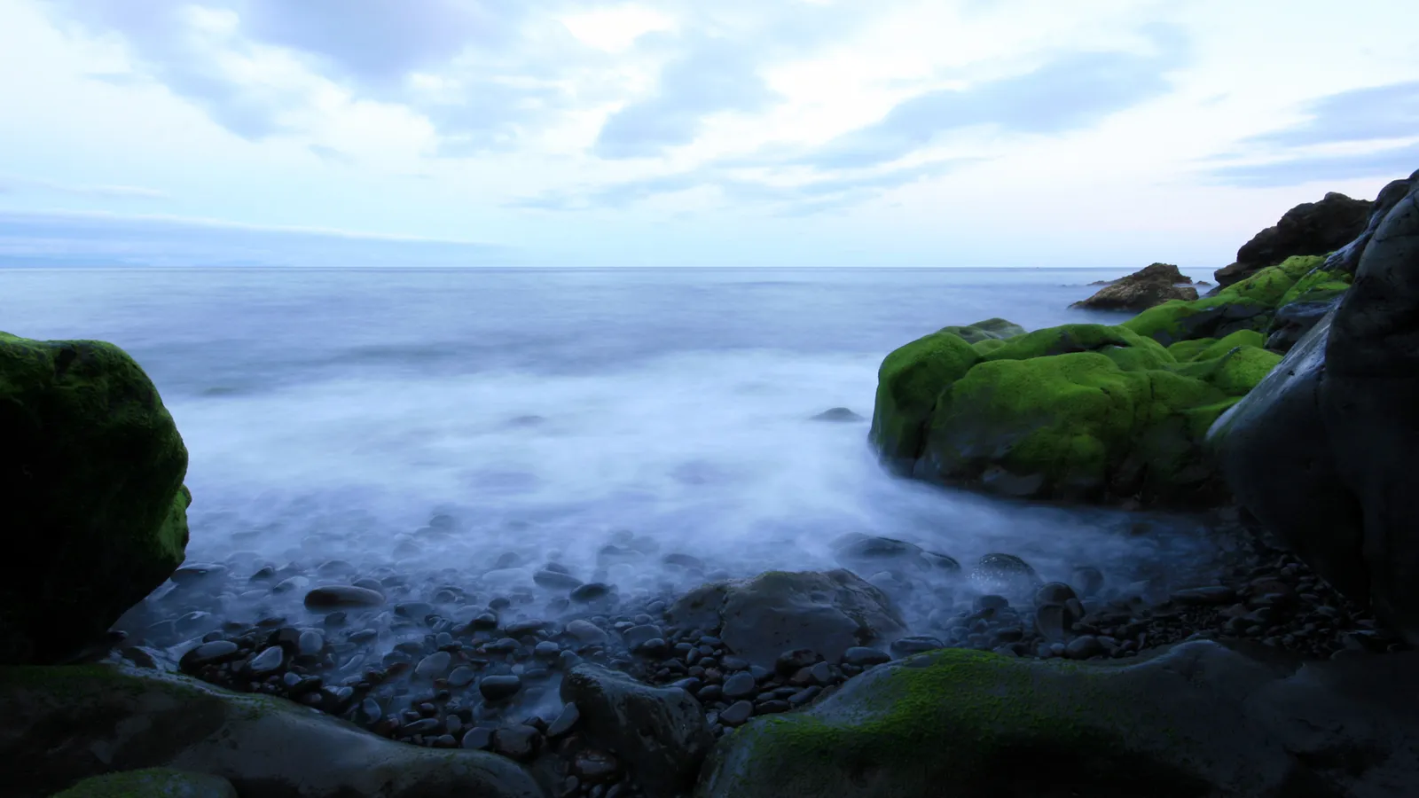 A rocky coastline with vibrant green moss on the rocks and the ocean waves creating a misty effect.