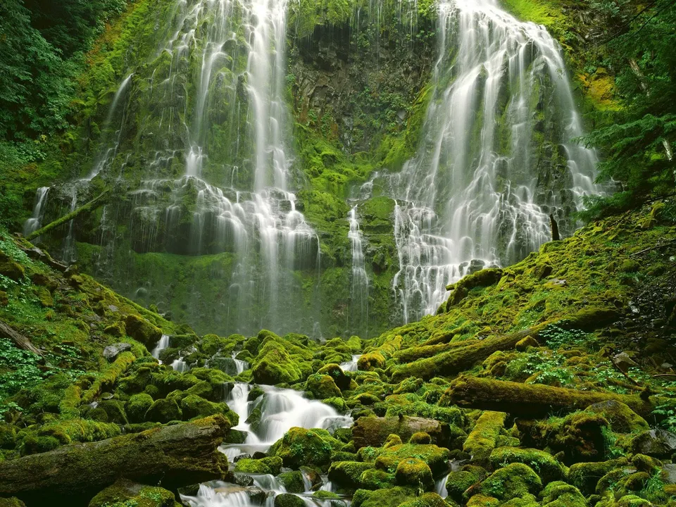 A wide, multi-tiered waterfall flows down a steep, moss-covered cliff face into a rocky stream below.