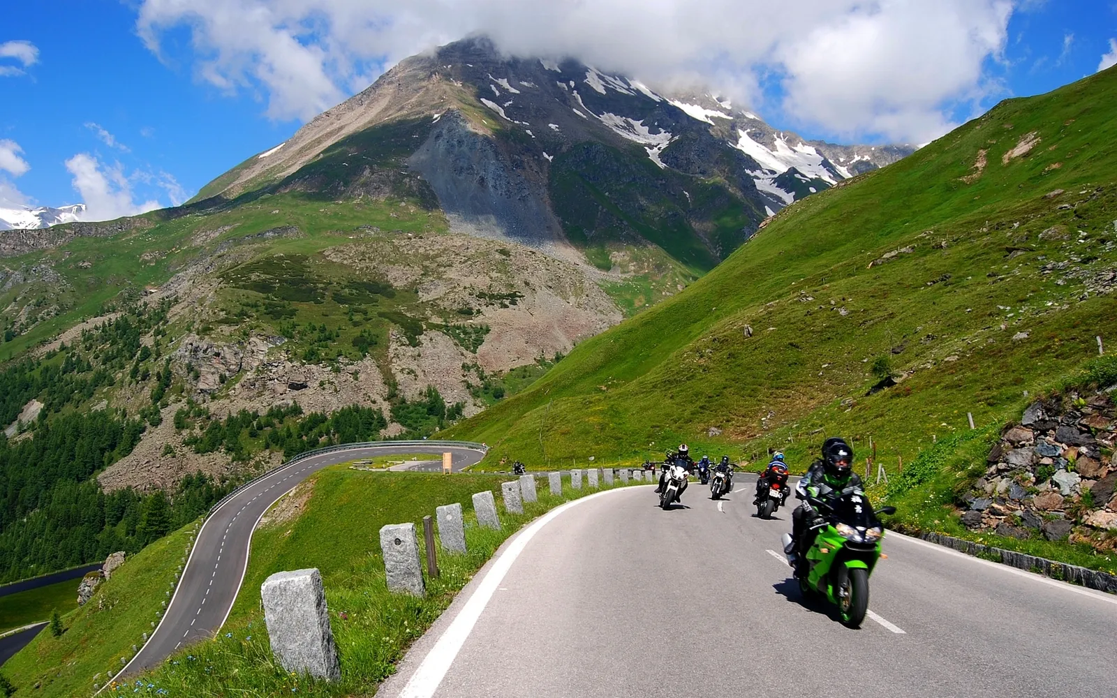 A group of motorcyclists ride along a winding mountain road with green hills and snow-capped peaks.