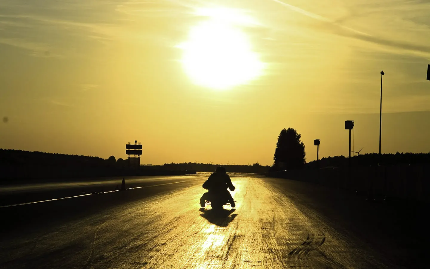 A lone motorcyclist is silhouetted against a bright, golden sunset sky on a racing track.