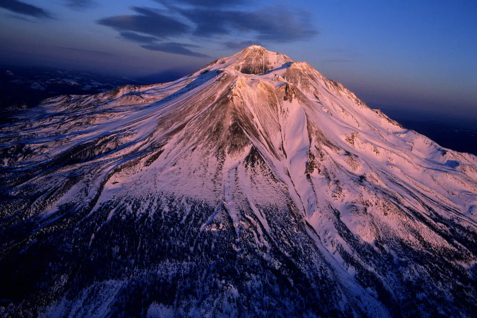 An aerial view of Mount Shasta covered in snow, illuminated by the soft light of sunrise.