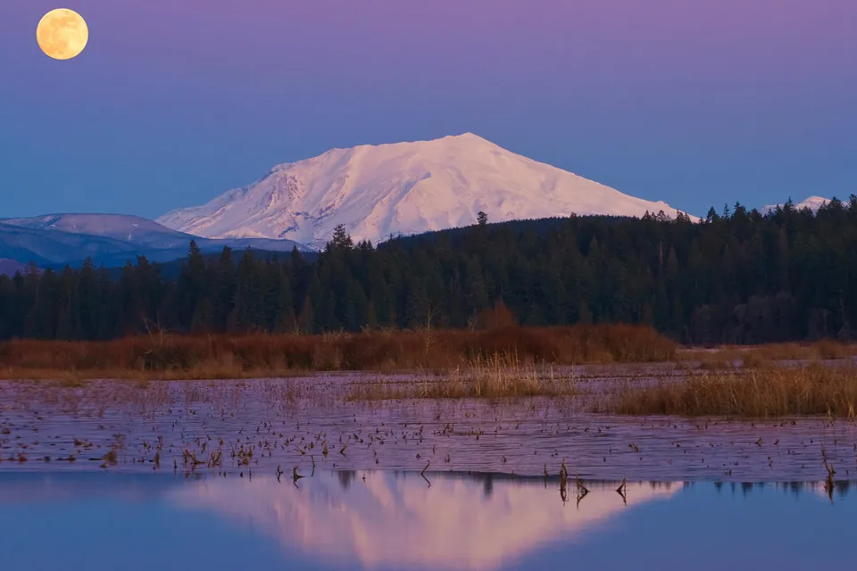 A snow-covered Mount St. Helens volcano under a large full moon with a reflective lake in the foreground.