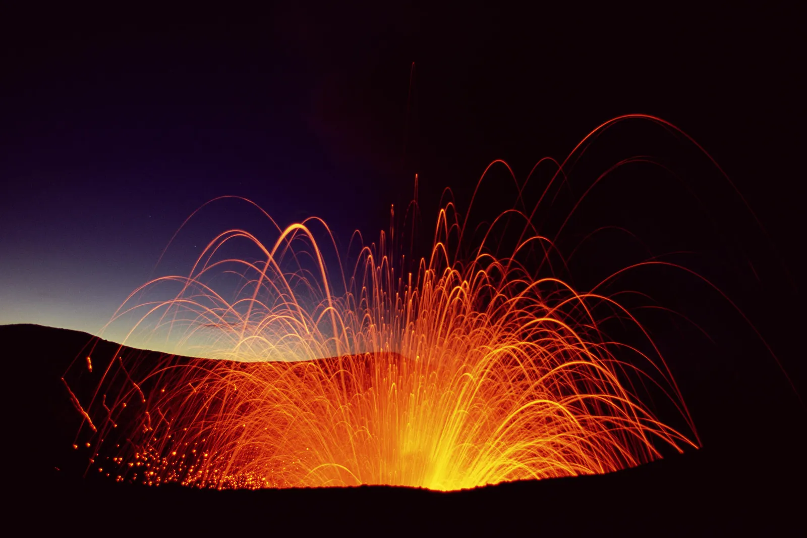 A volcano erupts at night, spewing glowing embers and lava trails into the dark sky.